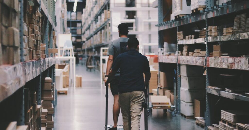 Two men maneuver a trolley in a large warehouse filled with boxes and shelves.