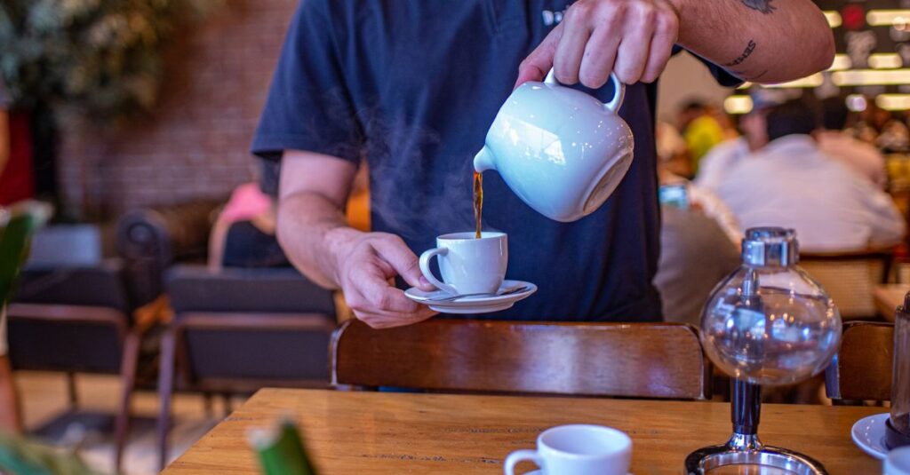 Barista expertly pouring hot tea into a white cup in a bustling cafe.