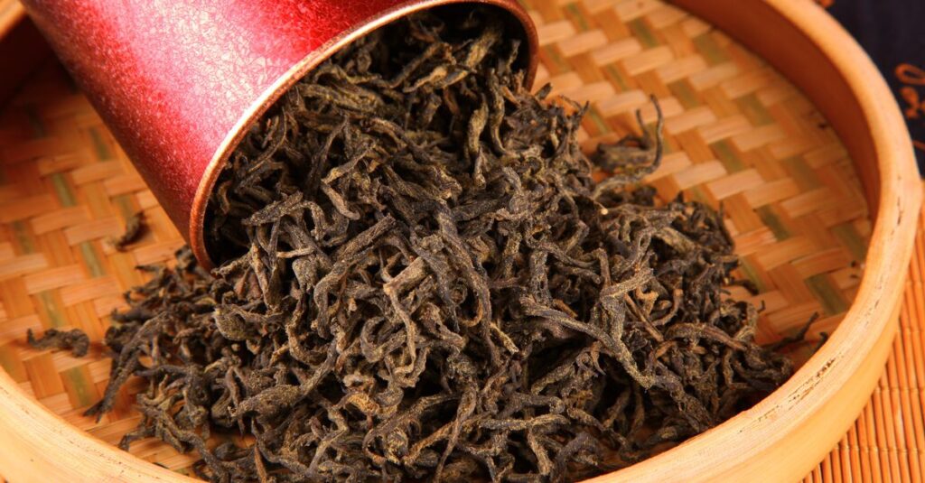 Close-up of loose black tea leaves spilling from a red canister onto a woven tray.