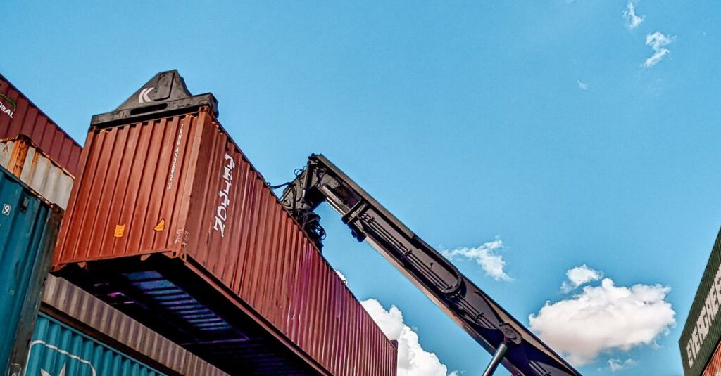 Red hydraulic crane handling shipping containers at a port under a clear blue sky.