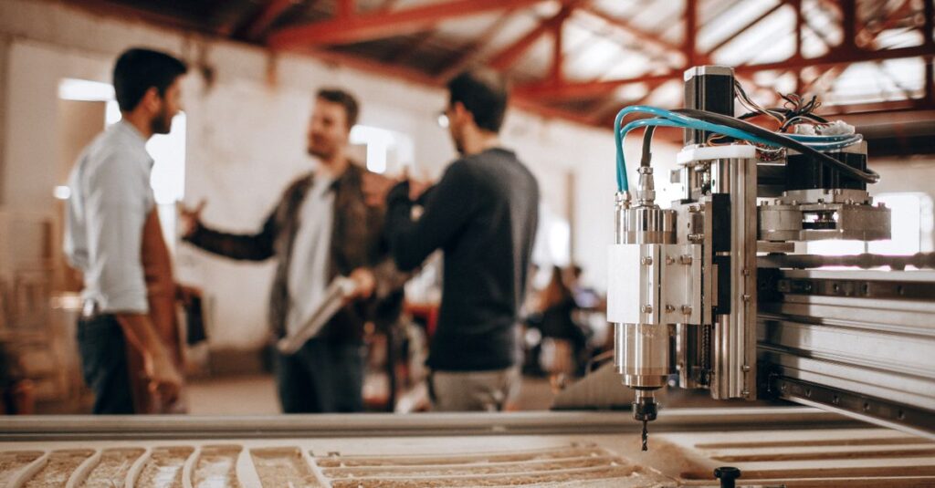 Three men discussing work in a woodworking shop with CNC machinery in focus.
