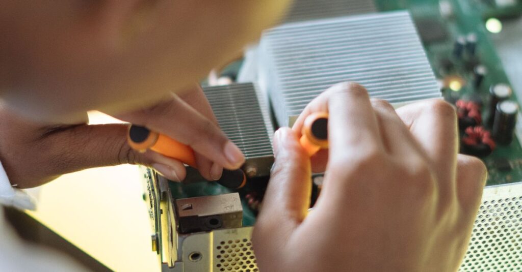 Crop of ethnic system administrator in uniform using screwdrivers and checking motherboard on electronic equipment during work in repair department