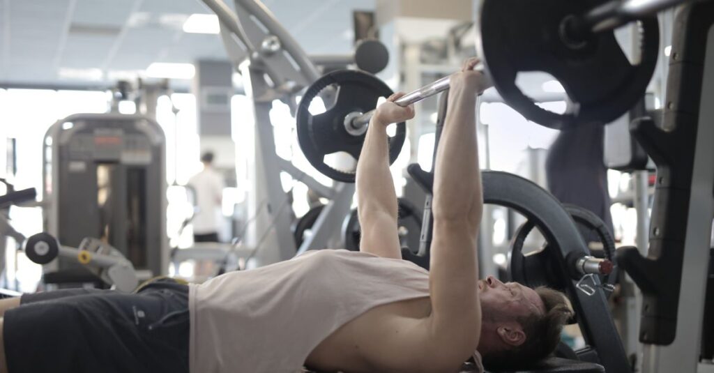 Side view of strong young sportsman in casual sportswear pressing heavy barbell on bench while intense training in modern fitness center