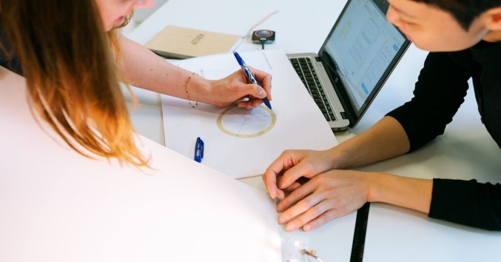 Two women collaborate on a design project at their desk, focusing on drawings and laptop.