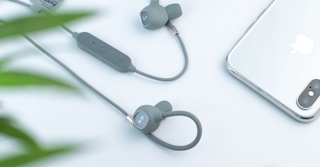 From above through green plant leaves arrangement of modern wired earphones and headphones placed on white desk near silver mobile phone