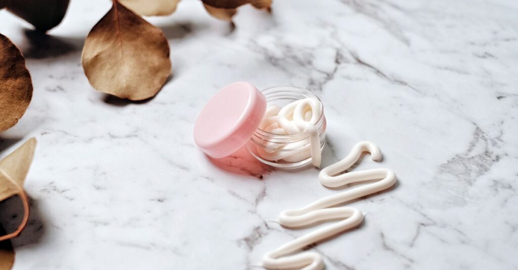 Close-up of skincare cream in a pink glass jar on a marble surface surrounded by leaves, ideal for beauty and wellness themes.