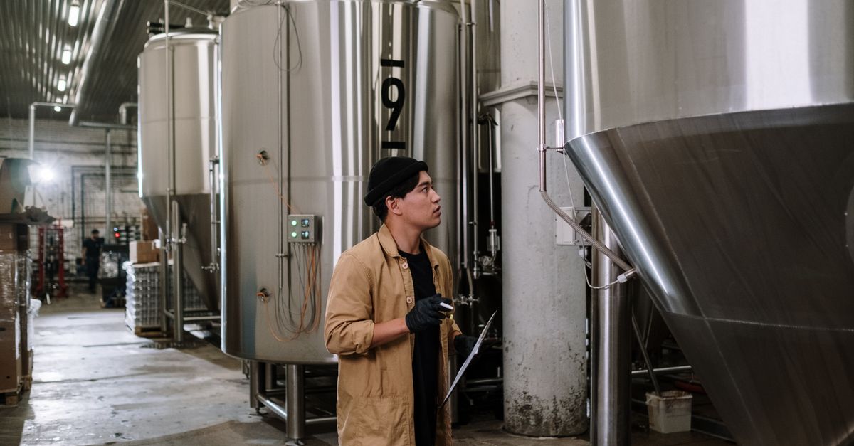Asian man inspecting stainless steel tanks in a brewery, showcasing modern industrial processes.