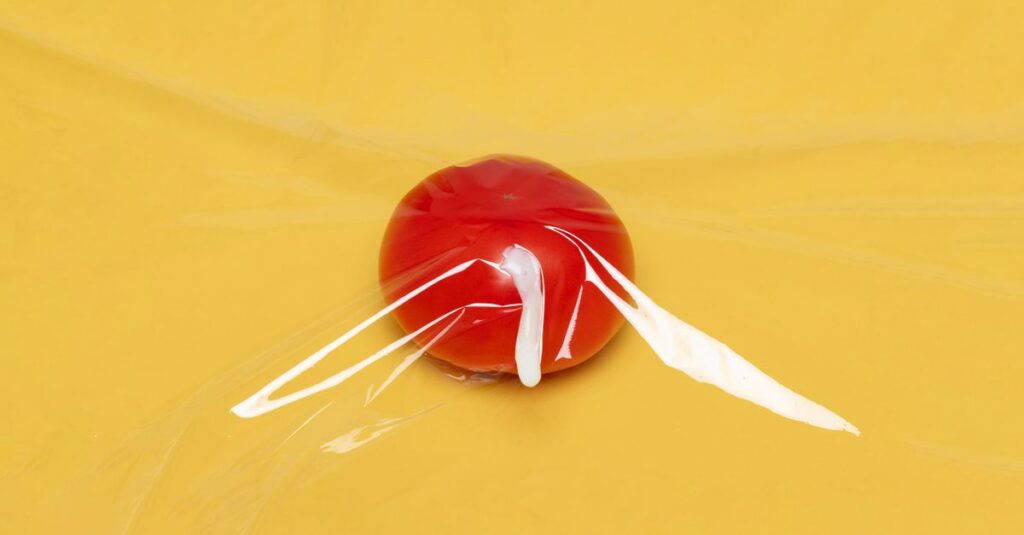A fresh tomato wrapped in clear plastic wrap on a vibrant yellow background, highlighting freshness and packaging.