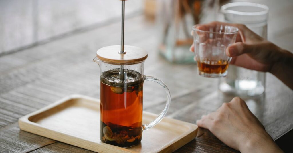 Crop anonymous female enjoying aromatic black tea and sitting at table with French press teapot on tray