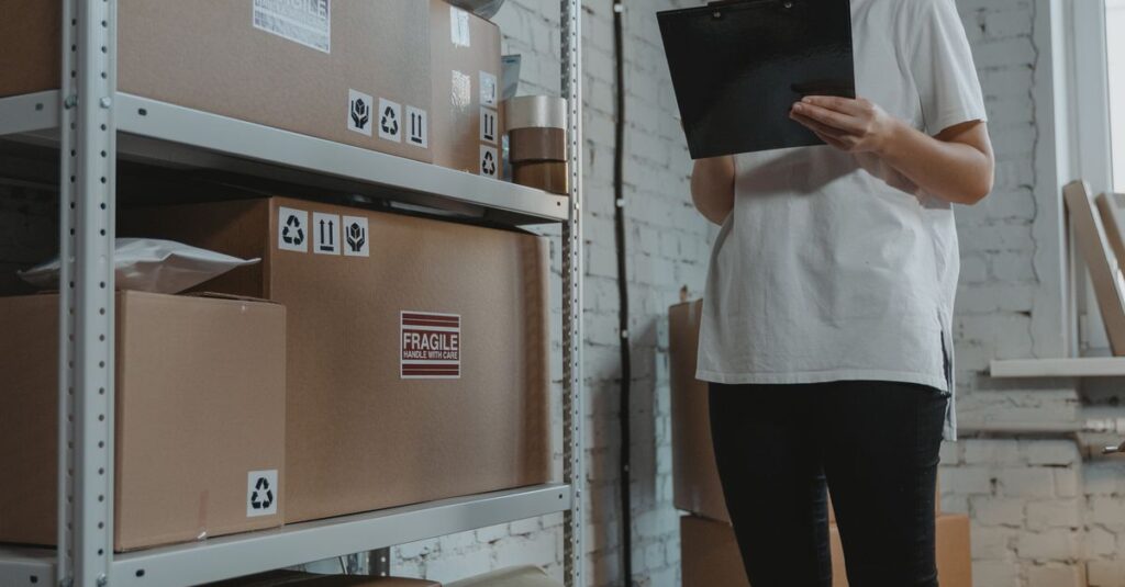 A female worker in casual attire organizes packages in a warehouse setting, examining a clipboard for inventory management.