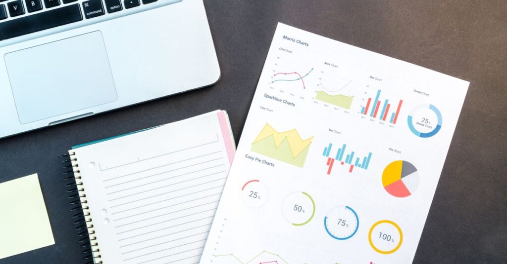 Flatlay of business report with colorful charts, a notebook, and a laptop for data analysis on a desk.