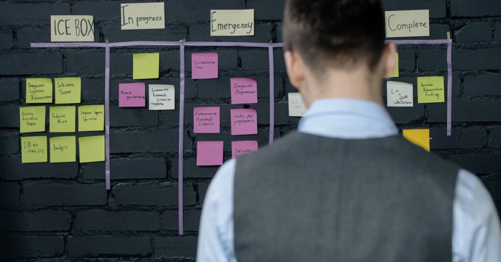 A person in a vest stands in front of a project management board with sticky notes on a dark brick wall.