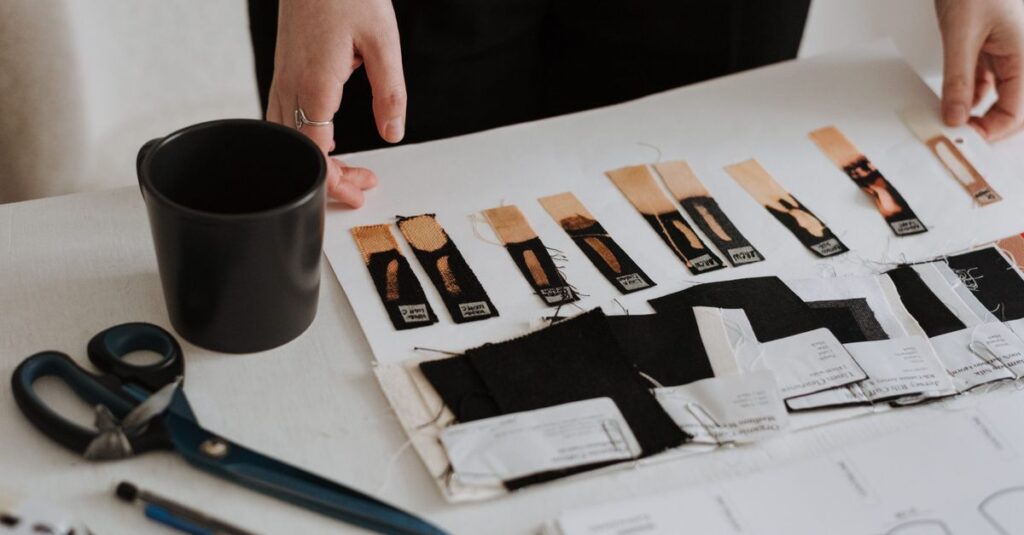 Unrecognizable designer standing at table with samples of various fabrics for new collection and scissors while working in light modern studio