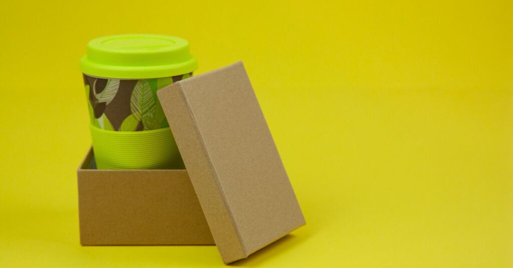 Composition of colorful paper cup with light green lid on it in small cardboard box on yellow surface