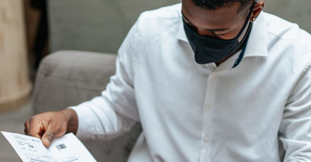 Adult man in a white shirt sits indoors, attentively examining documents while wearing a face mask.