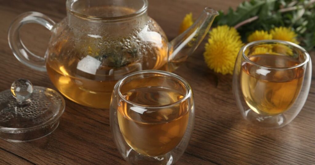 Close-up of herbal dandelion tea in glass teapot and cups on a wooden surface with flowers.