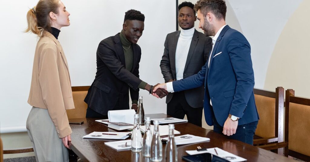 Four professionals in a modern office, two men shaking hands signifying agreement.